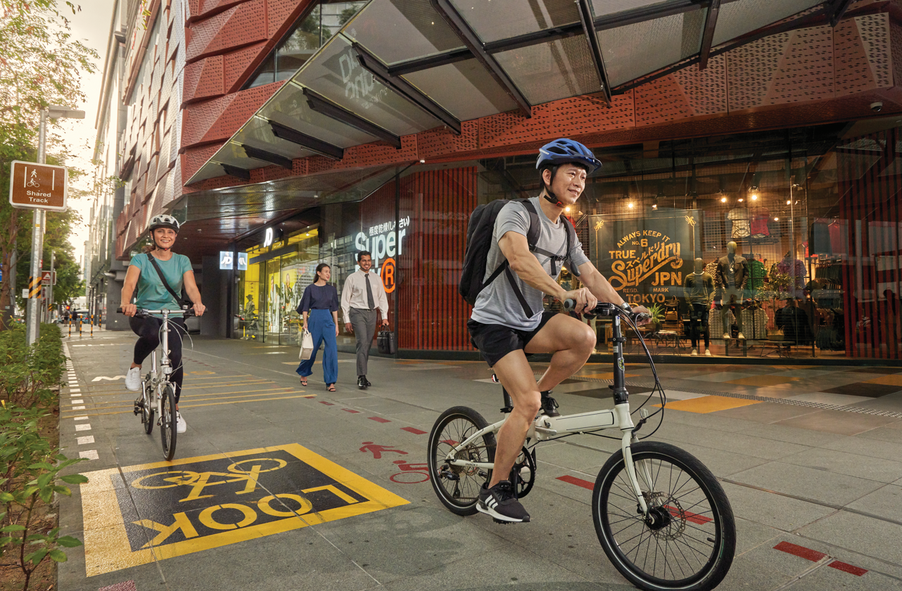 People cycling on a shared path in a city, near shops and pedestrians.